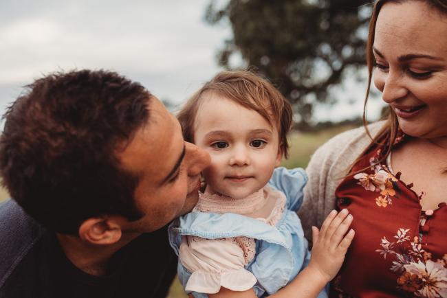 Father leaning in to kiss his daughter who is being held by her mother with Perth family photographer at Perry's Paddock