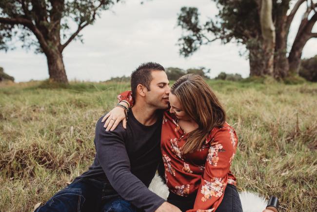 husband leaning over to kiss his wife as they sit on the grass with Perth family photographer at Perry's Paddock