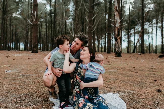 Family-Photographer-Perth-The-Pines-11-of-21 Mother holding her baby as she wipes the mouth of her older son and dad sits with them during a Family photographer session at The Pines in Wanneroo