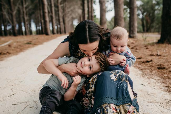 Mother holding her baby on her lap and kissing her little boy who has his head on her lap during a Perth family photography session at the Pines Wanneroo
