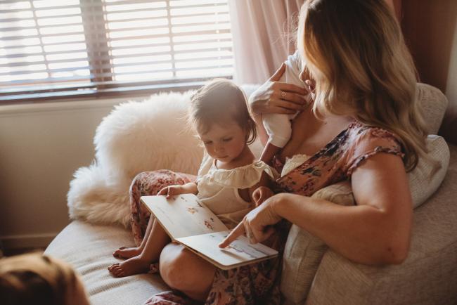 Mother sitting on a chair with her new baby over her shoulder and her daughter on her lap as she reads a story during an in-home Perth newborn lifestyle session