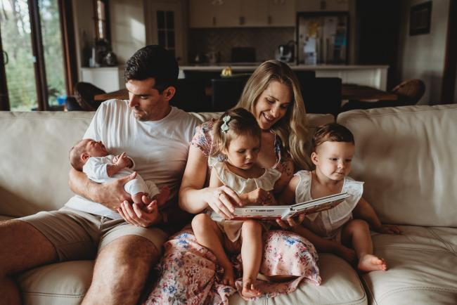 Family of 5 sitting on the couch as mum reads to the girls and dad holds their new baby during an in-home Perth newborn lifestyle session
