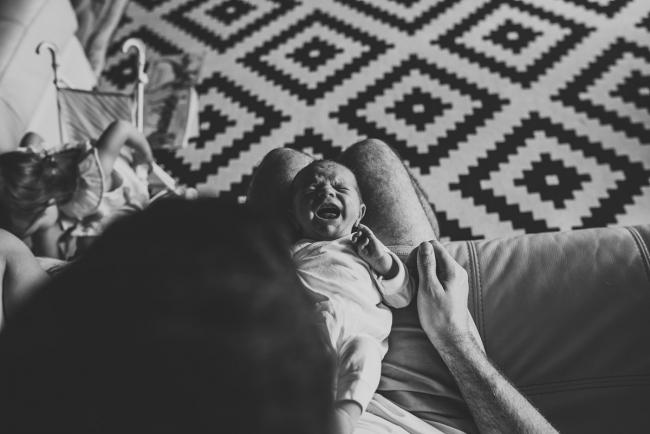 Black and white image of baby laying on his dad's lap and crying during an in-home Perth newborn lifestyle session