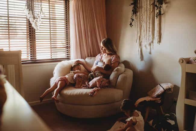 Mother sitting cross legged on a big round chair feeding her baby while touching her daughter's hair during an in-home Perth newborn lifestyle session