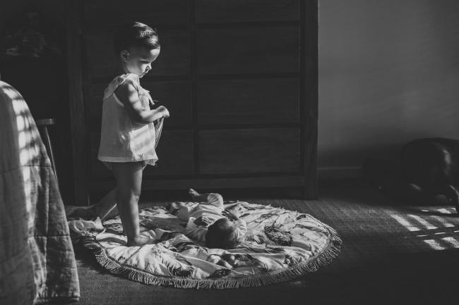 Black and white image of new baby laying on the floor as big sister stands next to him during an in-home Perth newborn lifestyle session