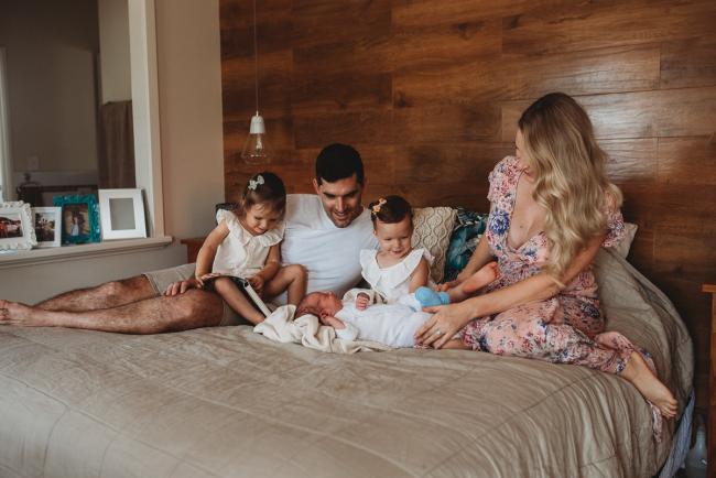 New baby laying on a bed with his parents and older two sisters looking at him during an in-home Perth newborn lifestyle session