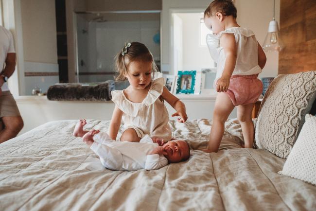 Baby laying on a bed as big sisters stand behind him during an in-home Perth newborn lifestyle session
