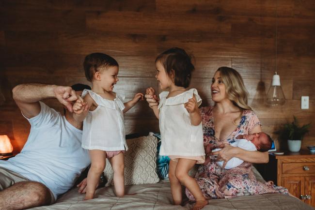 Little girls jump on the bed as parents hold their hands and mum holds their new baby during an in-home Perth newborn lifestyle session
