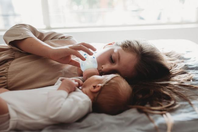 Little girl laying on the bed and feeding her baby sister a bottle during a Perth Newborn photography session