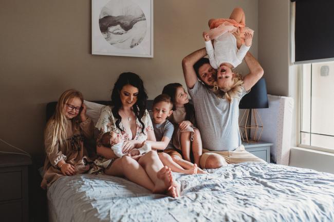 Family of 5 kids and parents on the bed, with father holding daughter upside down during a Perth Newborn photography session