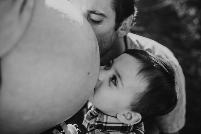 Black and white image of father and son kissing mother's pregnant tummy during a Perth maternity photography session at The Pines at golden hour