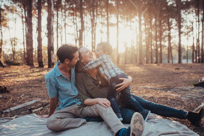 Parents sitting on a rug and little boy running up to hug mum during a Perth maternity photography session at The Pines at golden hour