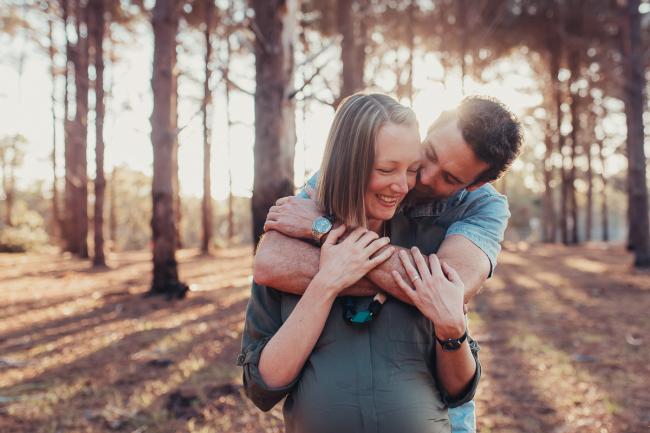 Husband and wife laughing as he hugs her from behind during a Perth maternity photography session at The Pines at golden hour