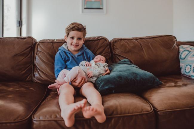 Newborn-Lifestyle-Photographer-Perth-26-of-54 Little boy smiling at the camera as he holds his new baby sister during a lifestyle newborn photography session in Perth