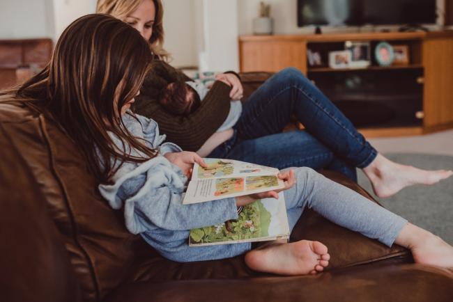 Newborn-Lifestyle-Photographer-Perth-3-of-54 Little girl sitting on the couch reading a book next to her mother who is breastfeeding during a lifestyle newborn photography session in Perth