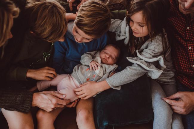 Newborn-Lifestyle-Photographer-Perth-30-of-54 Top down image of baby laying across the lap of her siblings during a lifestyle newborn photography session in Perth