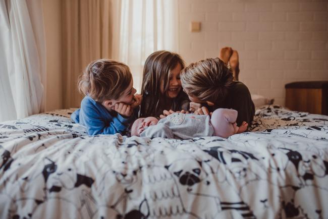 Newborn-Lifestyle-Photographer-Perth-33-of-54 Three older siblings laughing and looking at each other as their baby lays in front of them during a lifestyle newborn photography session in Perth