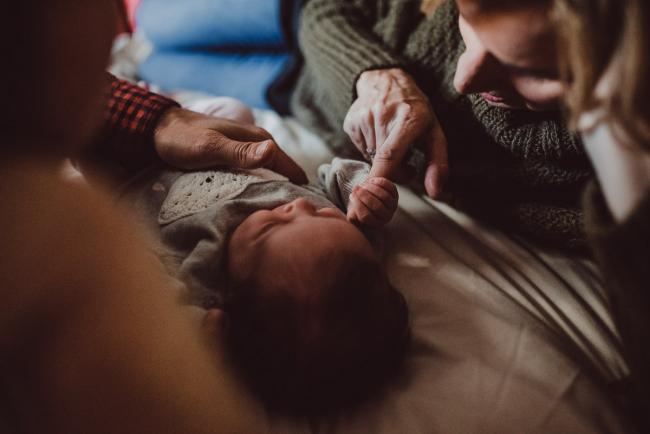 Newborn-Lifestyle-Photographer-Perth-45-of-54 Parents laying on the bed with their baby and mother holding baby's hand during a lifestyle newborn photography session in Perth
