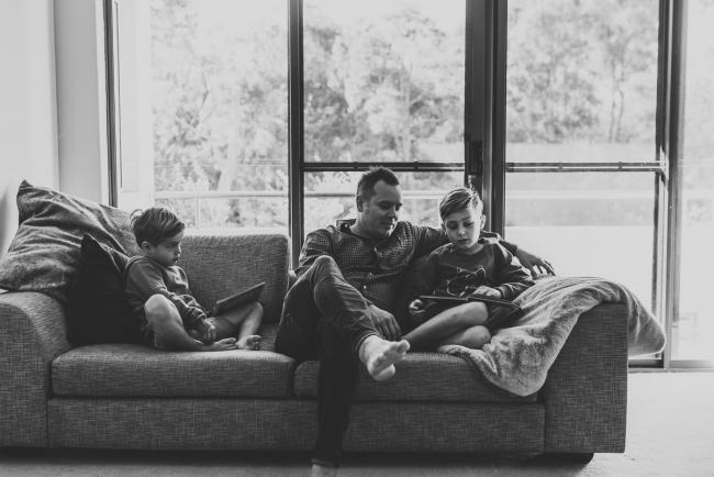 Newborn-Lifestyle-Photographer-Perth-5-of-54 Black and white image of father and two sons sitting on the couch during a lifestyle newborn photography session in Perth