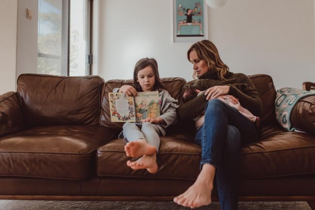 Newborn-Lifestyle-Photographer-Perth-7-of-54 Little girl reading on the couch next to her mother who is breastfeeding during a lifestyle newborn photography session in Perth