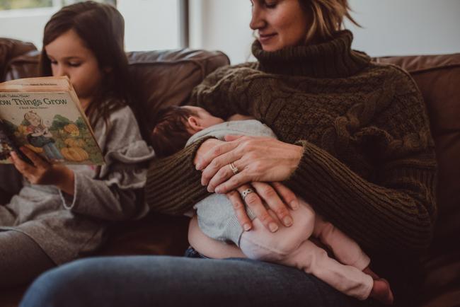 Newborn-Lifestyle-Photographer-Perth-8-of-54 Mother breastfeeding her baby as her older daughter reads next to her during a lifestyle newborn photography session in Perth