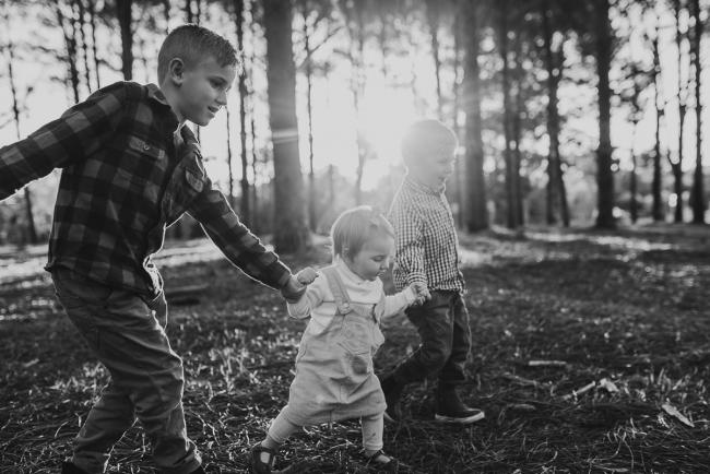 The-Pines-Family-Photographer-Perth-16-of-60 Black and white image of three siblings holding hands and walking during a Perth Family Photography session during Golden Hour at The Pines