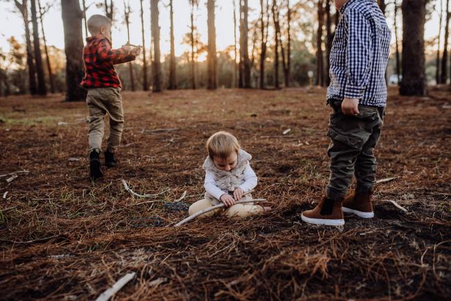 The-Pines-Family-Photographer-Perth-49-of-60 Little girl playing with a stick while her older brothers run around her during a Perth Family Photography session during Golden Hour at The Pines
