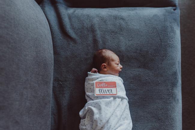 Top down image of wrapped baby laying on the couch with a 'hello my name is Oliver' sign during a Perth Fresh 48 photography session captured in home
