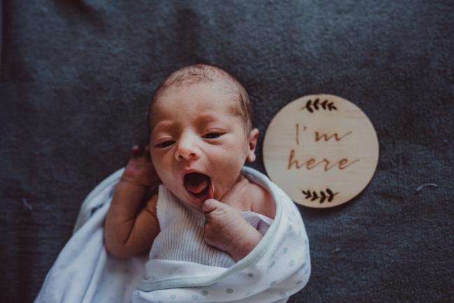 New baby pulling his lip down next to a wooden 'i'm here' sign during a Perth Fresh 48 photography session captured in home