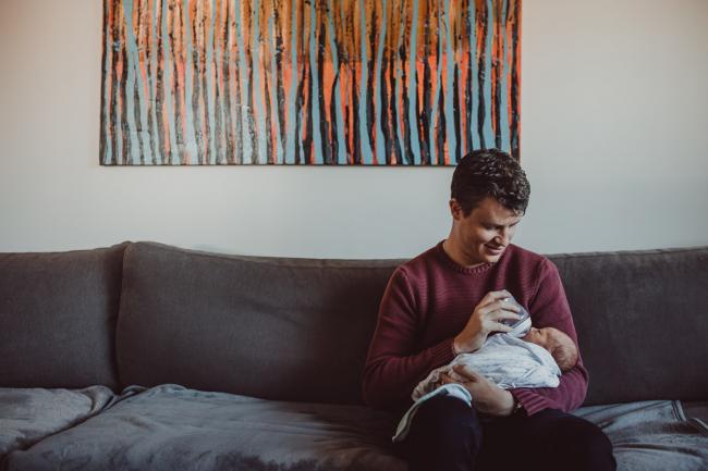 Father sitting on the couch bottle feeding his new baby during a Perth Fresh 48 photography session captured in home