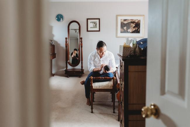 Looking through the doorwat as father holds his baby and mother's reflection is in the mirror during an in-home Perth Newborn Lifestyle photography session