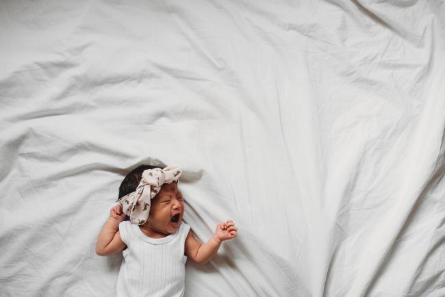 New baby laying on the bed crying with a bow in her hair during an in-home Perth Newborn Lifestyle photography session