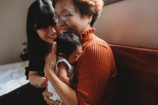 Grandmother holds her granddaughter as her daughter smiles next to her during an in-home Perth Newborn Lifestyle photography session
