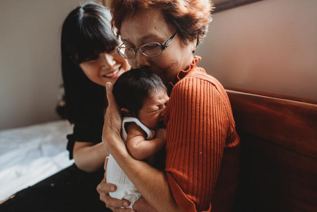 Grandmother kisses granddaughter as her daughter sits next to her during an in-home Perth Newborn Lifestyle photography session