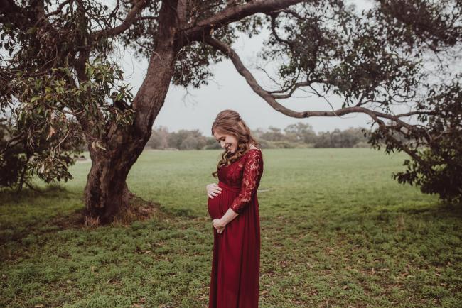 Perth-Maternity-Photographer-Perrys-Paddock-5-of-39 Lady in a red dress standing under a tree and looking at her baby bump during a Perth maternity session at Perry's Paddock during rain