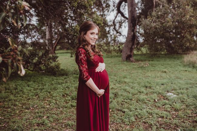 Perth-Maternity-Photographer-Perrys-Paddock-6-of-39 Lady in red dress standing sideways and smiling at the camera during a Perth maternity session at Perry's Paddock during rain