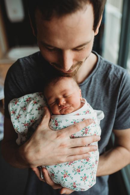 Father holding his wrapped baby against his chest as he smiles down at her during a Perth Fresh 48 photography session at SJOG Mt Lawley