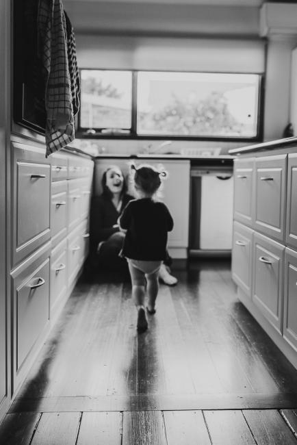 black and white image of little girl running to her mother who is sitting on the floor in the kitchen with Perth lifestyle photographer
