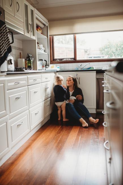 mother sitting on the floor in her kitchen holding her new baby and talking to her toddler with lifestyle newborn photographer Perth