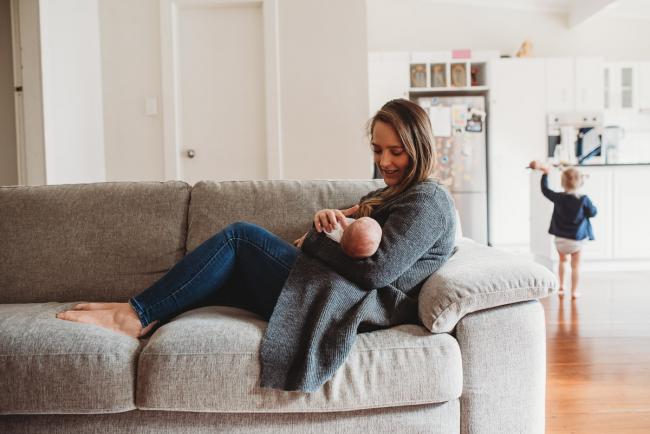 mother feeding her new baby on the couch with toddler daughter in the background with lifestyle newborn photographer Perth