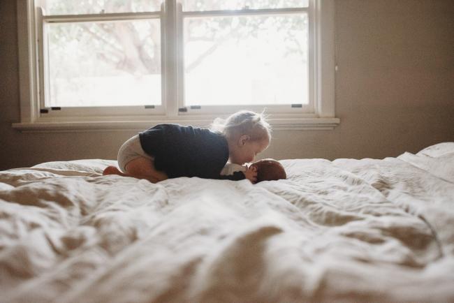 Toddler sister bending over and kissing her new baby brother on the bed with lifestyle newborn photographer Perth