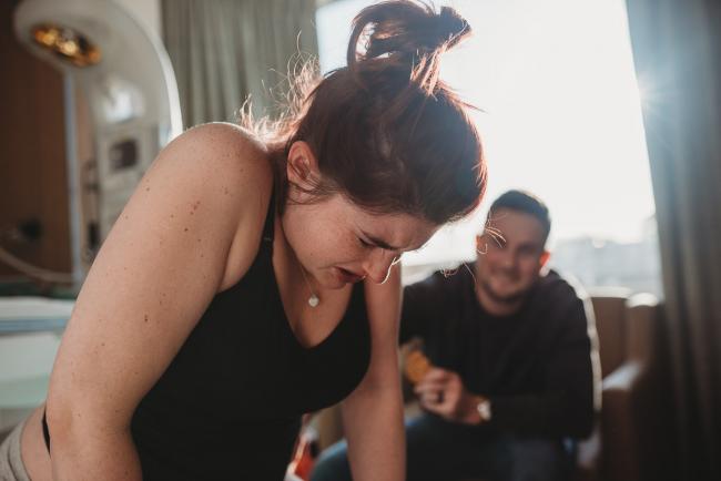 Birth-Photographer-Perth-18-of-60 Pregnant woman leaning over during contraction with partner in the background with Perth Birth Photographer at SJOG Mt Lawley