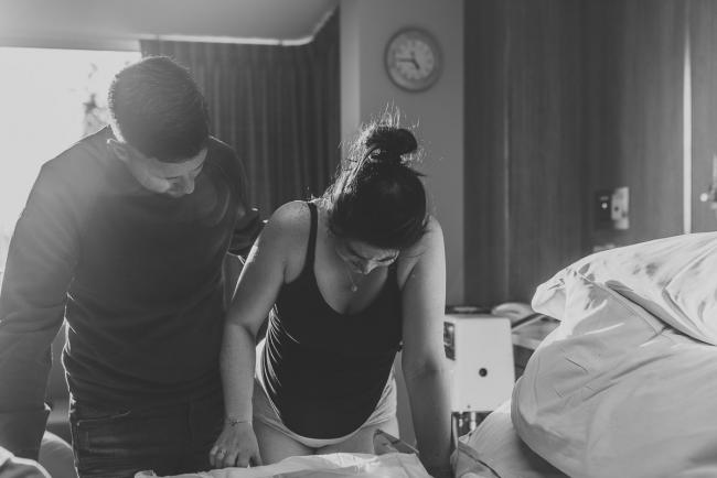 Birth-Photographer-Perth-22-of-60 Black and white image of pregnant woman leaning over hospital bed with partner rubbing her back with Perth Birth Photographer at SJOG Mt Lawley