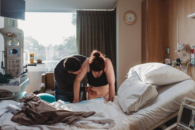 Birth-Photographer-Perth-23-of-60 Labouring mother leaning over bed with partner by her side with Perth Birth Photographer at SJOG Mt Lawley