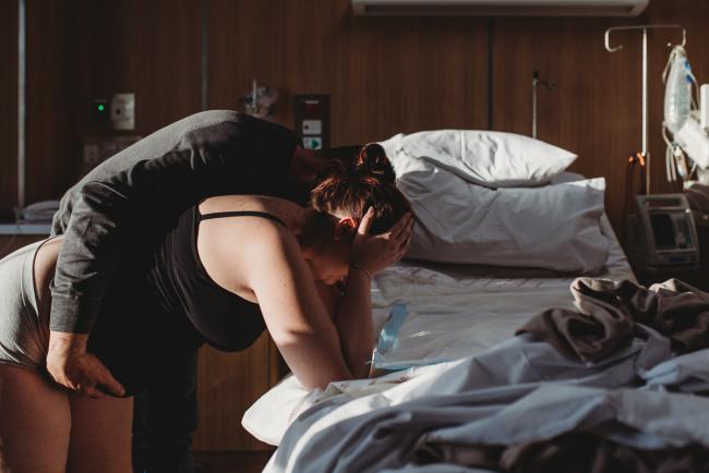 Pregnant woman leaning over hospital bed during labour with partner hugging her with Birth Photographer Perth at SJOG Mt Lawley