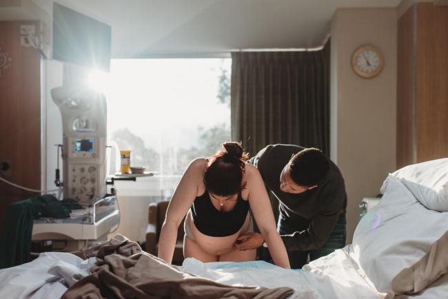 Pregnant woman leaning over bed during contraction with partner next to her with Birth Photographer Perth at SJOG Mt Lawley