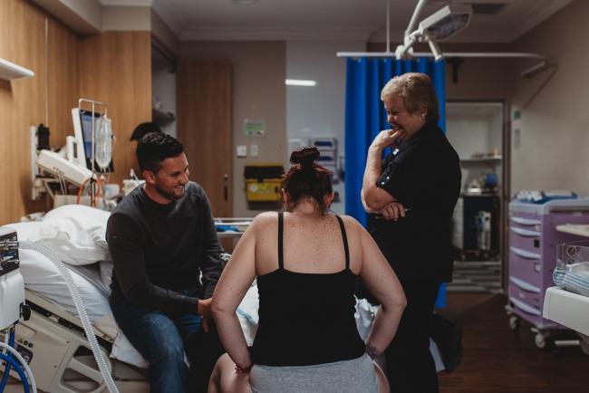 Birth-Photographer-Perth-34-of-60 Labouring mother on fit ball with partner on bed and midwife next to her with Perth Birth Photographer at SJOG Mt Lawley