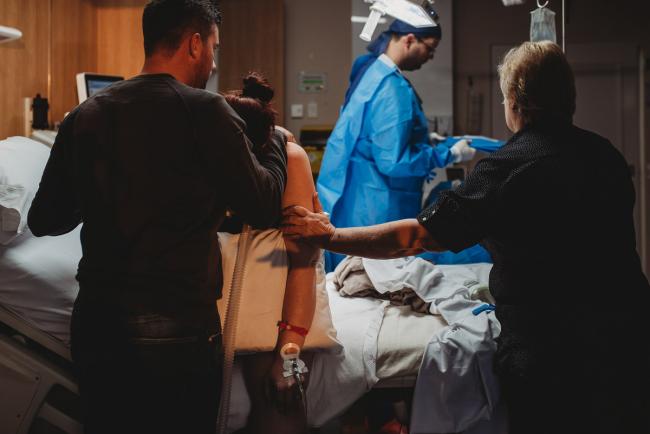 Pregnant woman on hospital bed with partner in front of her and midwife holding her arm with Birth Photographer Perth at SJOG Mt Lawley
