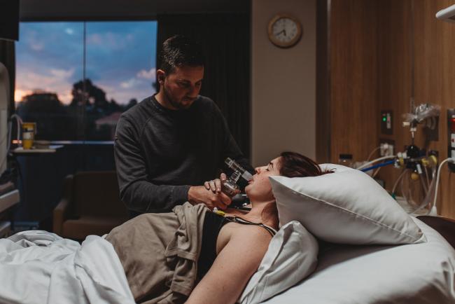 Birth-Photographer-Perth-44-of-60 Pregnant woman using gas on bed with partner by her side and sunset out the window with Perth Birth Photographer at SJOG Mt Lawley