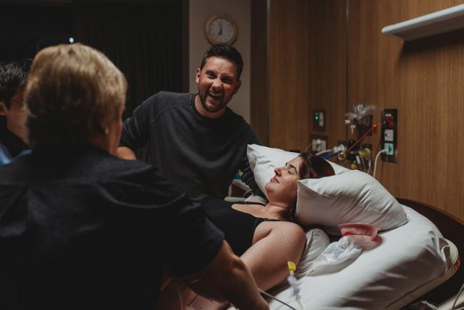 Birth-Photographer-Perth-53-of-60 Man laughing at the camera next to his pregnant partner in hospital bed with Perth Birth Photographer at SJOG Mt Lawley